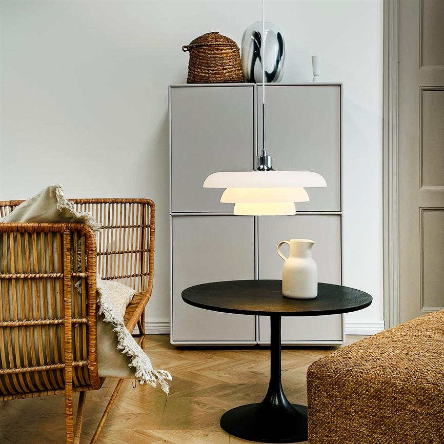 Hvid pendellampe hænger og lyser over et rundt sort sidebord med en hvid kande; i et lyst stuehjørne med rattanstol, skab, kurv og sildebensparketgulv.