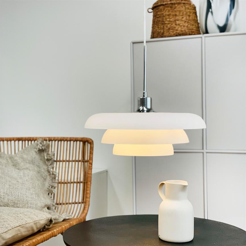 Hængende hvid pendellampe lyser varmt over et rundt sort bord; en hvid keramikkande står på bordet, med rottingstol med pude og hvide skabe i et minimalistisk rum.