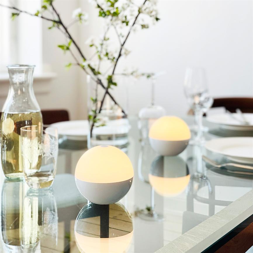 To runde bordlamper lyser blødt og varmt på et glasbord omgivet af karafler, glas, hvide tallerkener og en vase med blomstrende kvister i et lyst spiserum.