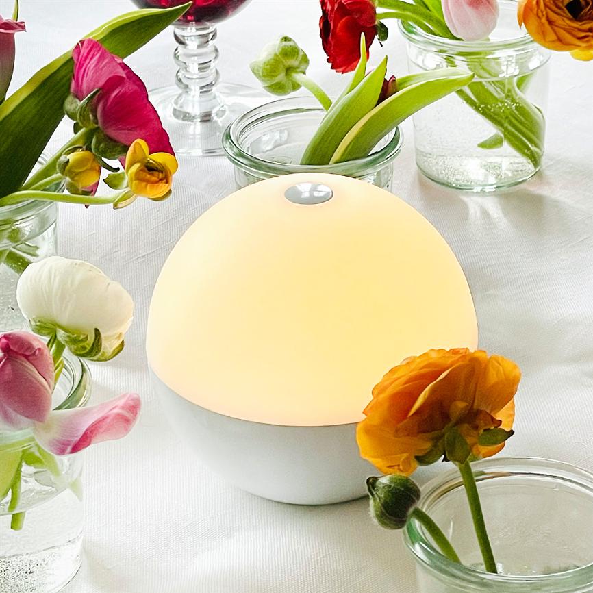 En rund, varmglødende bordlampe gløder blødt, omgivet af små glasvaser med farverige ranunkler og tulipaner på en hvid dug.