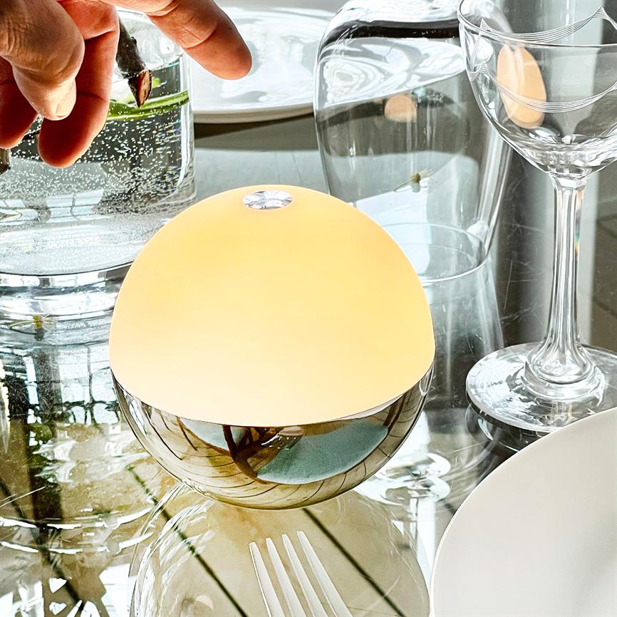 En rund bordlampe lyser blødt, dens krombund reflekterer omgivelserne; den står på et glasbord ved et vinglas, en gaffel og et glas med en plante, mens en hånd nærmer sig.