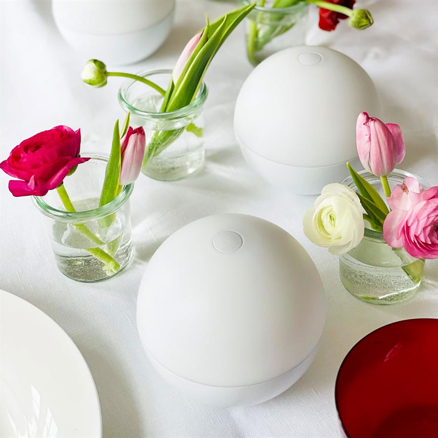 To hvide kugleformede enheder står placeret på en hvid dug, omgivet af små glasvaser med pink og hvide tulipaner og ranunkler, samt en hvid tallerken og et rødt glas.