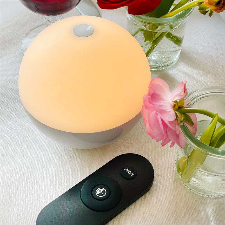 En rund bordlampe udsender varmt, dæmpet lys; står på en hvid dug blandt små glasvaser med pink og røde blomster, og en sort fjernbetjening ligger foran. 
 Tekst i billedet: "ON/OFF"