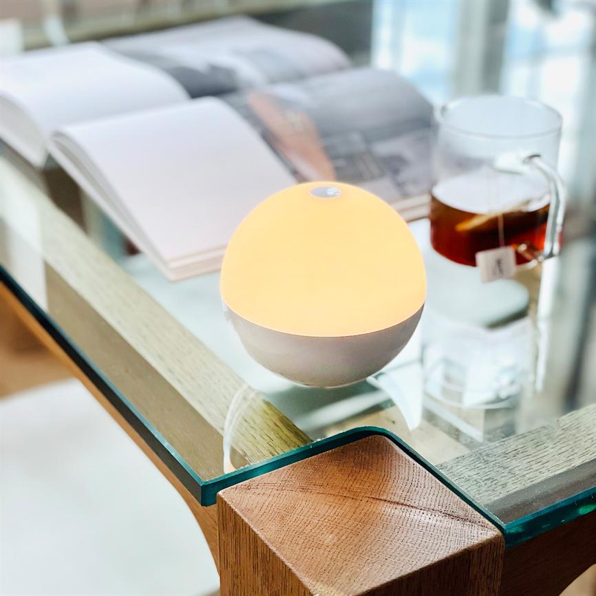 Lille rund lampe udsender varmt, blødt lys; står på et glasbord i hjørnet af et træbord, omgivet af et åbent magasin og en glaskop med te.
