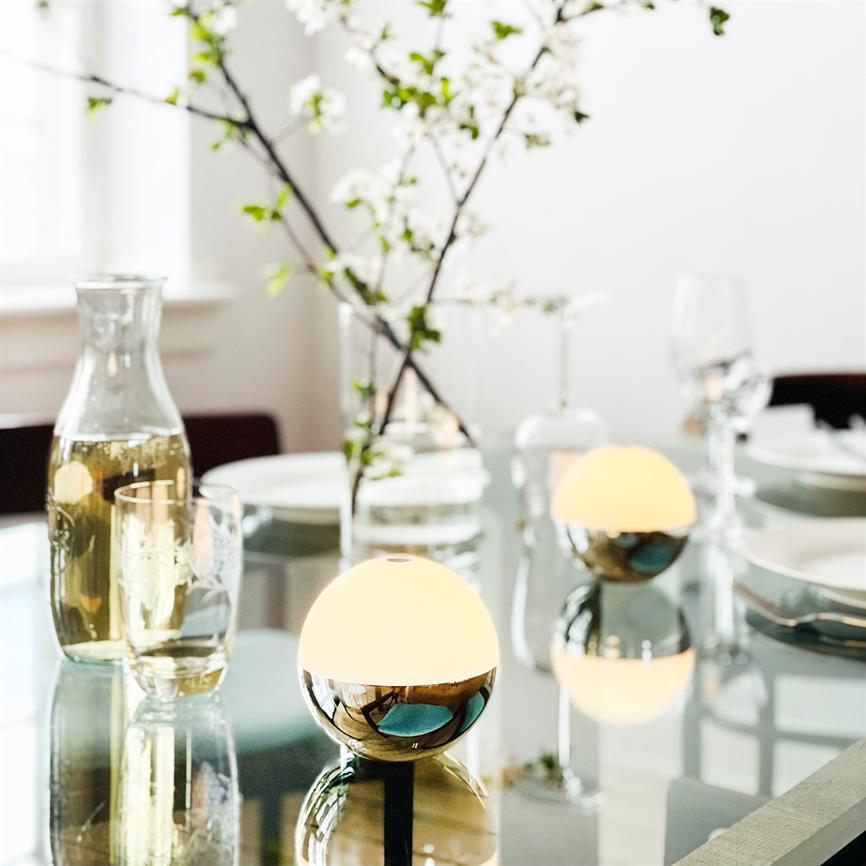 To små kugleformede bordlamper udsender varmt, blødt lys; de reflekteres i et glasbord omgivet af vinglas, en vandkaraffel, tallerkener og en vase med hvide blomster i en lys spisestue.