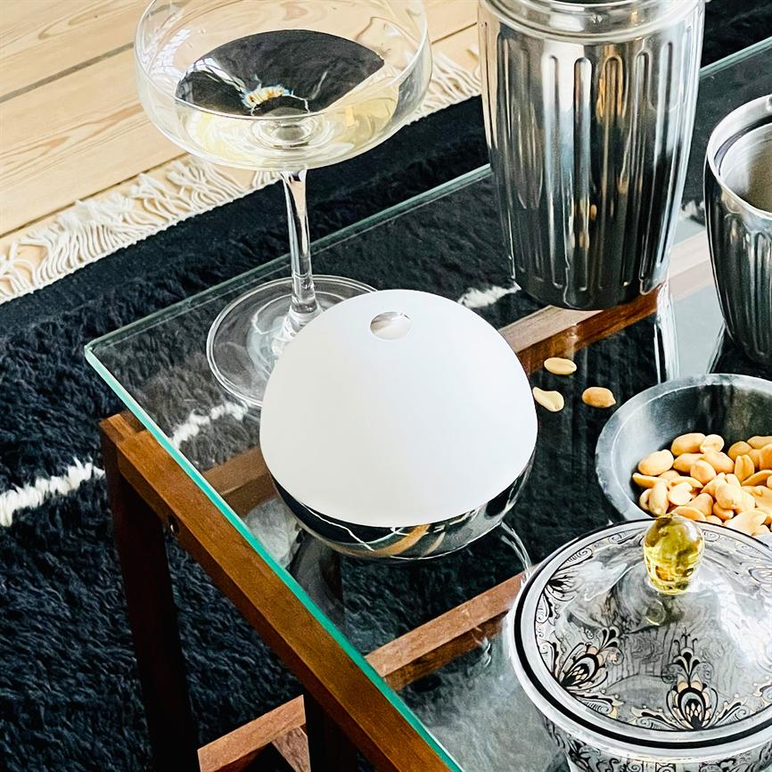 Hvid, halvkugleformet diffuser står på et glasbord, omkranset af et vinglas, metalbeholdere og en skål med peanuts på et sort, pelslignende tæppe.