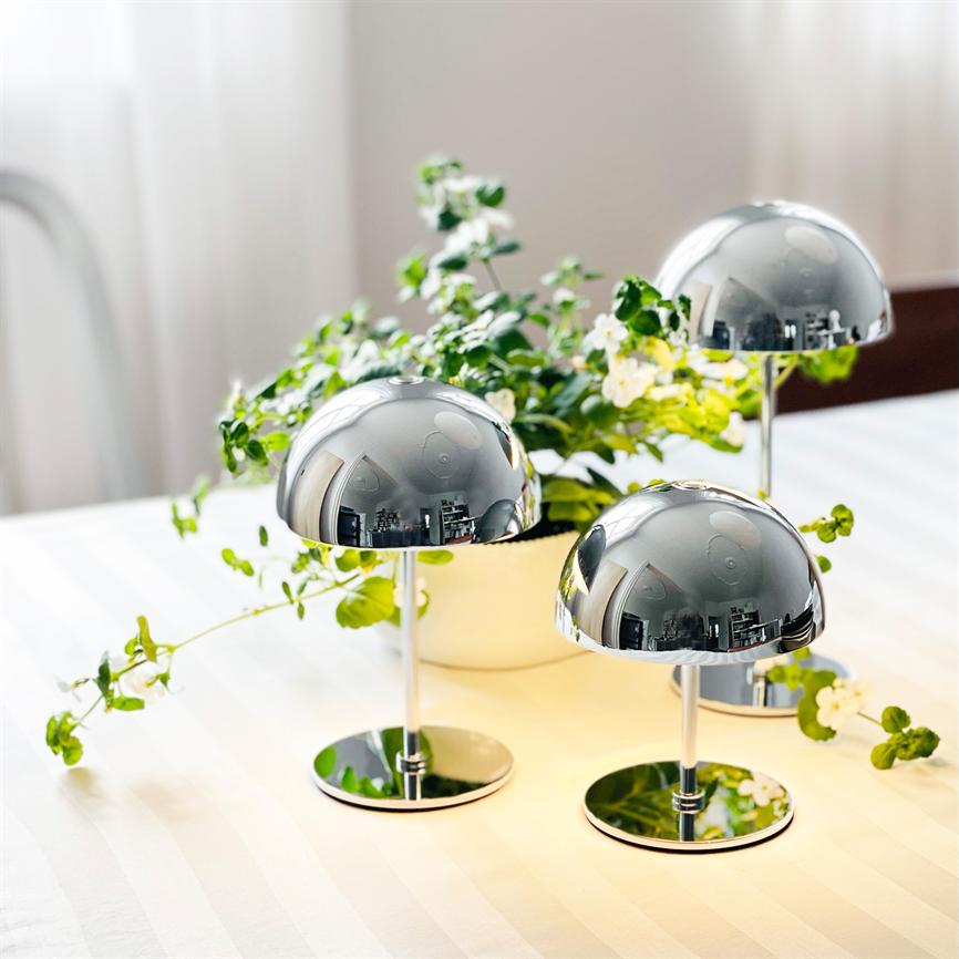Tre krombelagte svampelamper står på bordet og reflekterer rummet; foran en krukke med grøn plante og små hvide blomster på en stribet hvid dug i blødt dagslys.