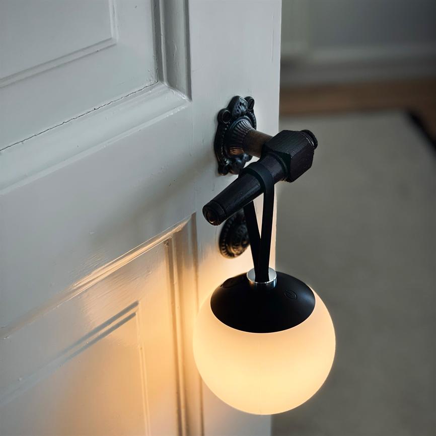 En kugleformet lampe hænger i en rem fra et ornamenteret dørgreb og kaster varmt, blødt lys på en hvid fyldningsdør i en dæmpet, tæppebelagt gang.