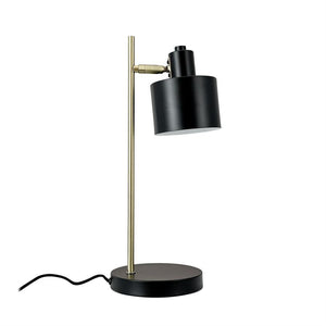 Bordlampe med sort cylindrisk skærm, vippet nedad, monteret på en slank messingstang og rund sort base; synlig sort ledning på ensartet hvid baggrund.