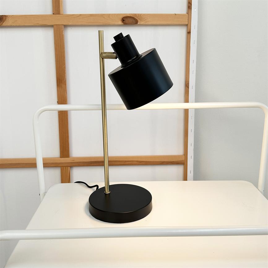 Sort bordlampe med messingstang og rund skærm vinklet nedad; står på en hvid metalhylde med det sorte kabel bagved, foran en treramme og hvid væg.