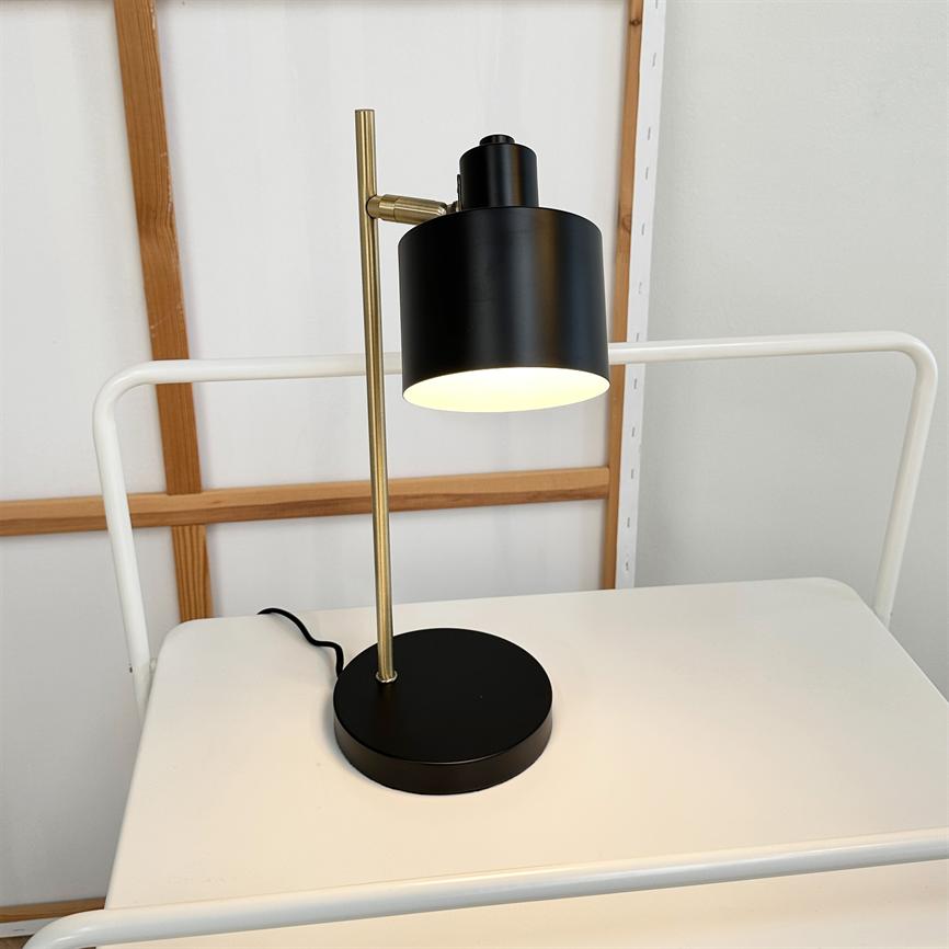 Sort og messing bordlampe lyser; cylindrisk skærm hænger fra en messingstang og står på rund sort fod på et hvidt bord foran en hvid væg med træ-lister.