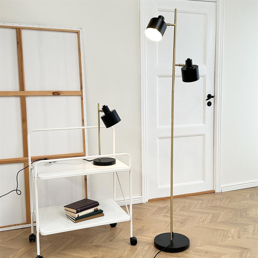 En høj gulvlampe med to sorte skærme lyser øverste skærm, stående ved en hvid dør på lyst sildebensparket; ved siden af et hvidt rullevognsbord med bordlampe, bøger og en tom lærredsramme.