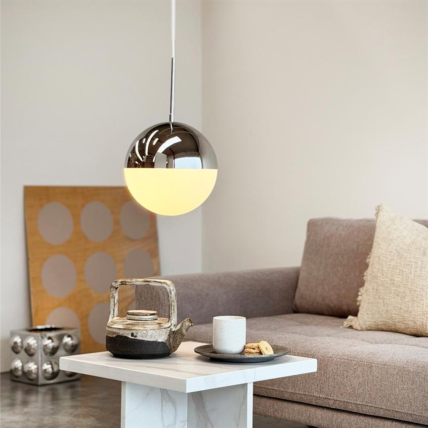 En rund pendellampe hænger tændt over et lille marmorbord; på bordet står en rustik tekande, kop og småkager. I baggrunden en beige sofa, pyntepude og dekorativt panel.
