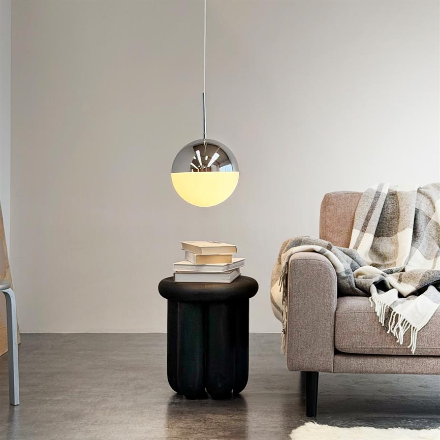 En rund hængelampe lyser over et lille sort sidebord med en stak bøger; ved siden af står en grå sofa med ternet plaid i et minimalistisk, lyst stuemiljø.