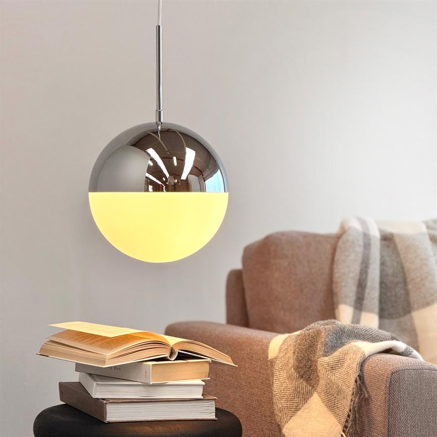 En rund pendellampe hænger og kaster blødt, varmt lys; under den står en stak åbne bøger på et bord ved en beige sofa med ternet plaid i et lyst rum.
