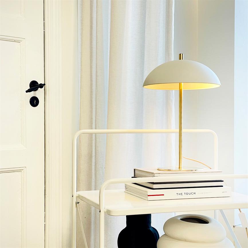 Bordlampe står tændt på en hvid hylde med bøger, én bog mærket "THE TOUCH", foran lyse gardiner og en lukket hvid dør. 
 Tekst: THE TOUCH