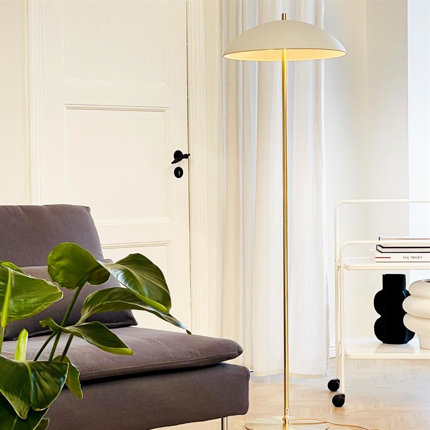 Gulvlampe står tændt og kaster varmt lys i en rolig stuemiljø ved en grå sofa, grøn stueplante, hvid dør og rullebord med bog og vaser. Tekst: "THE TOUCH".