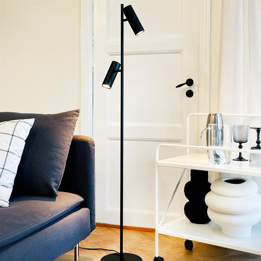 En slank, sort gulvlampe lyser ned ved en hvid dør, mellem en blågrå sofa med mønstret pude og en hvid rullevogn med glas, kande og dekorative vaser.