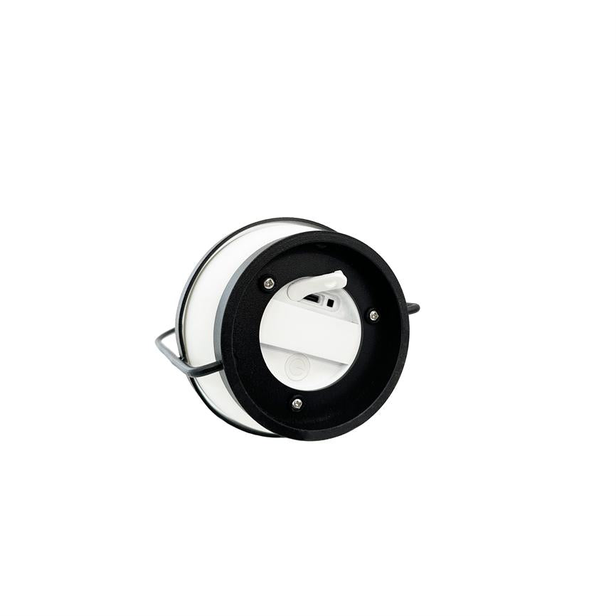 Rund bærbar LED-lampe ligger på siden; hvid kuppel og sort ydre ramme med synlige skruer. En foldbar metalbøjle understøtter den mod en neutral, hvid baggrund.