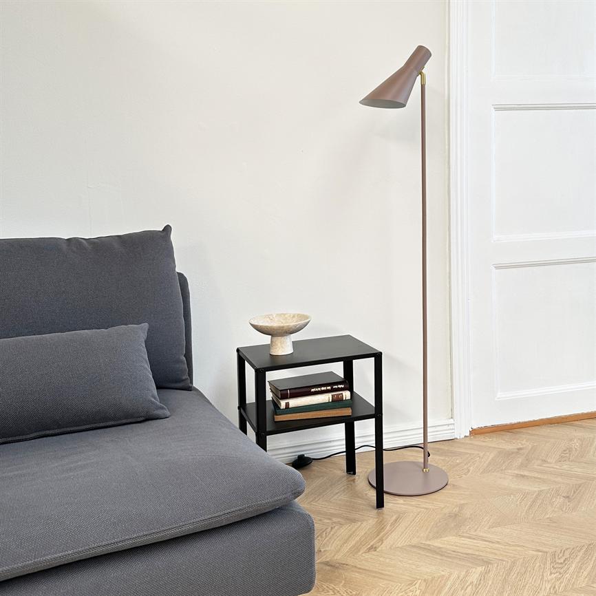 Grå sofa er placeret til venstre; et lille sort sidebord bærer en skål og en stak bøger; en slank gulvlampe står ved siden af i et lyst rum med parketgulv.