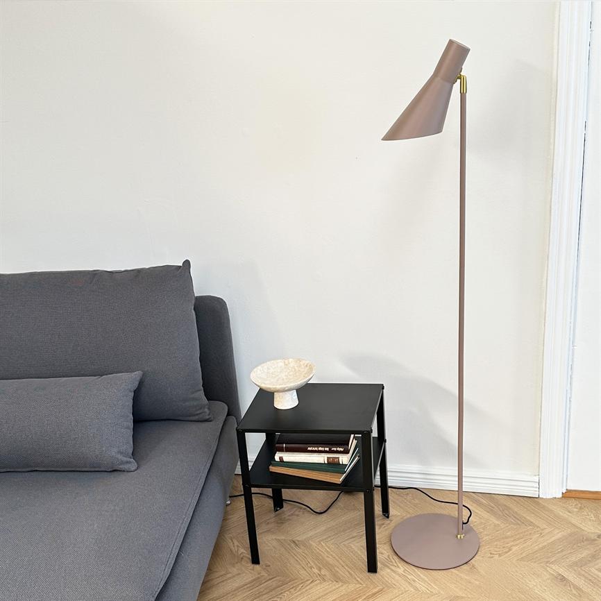 Gulvlampe står ved siden af et lille sort sidebord; bordet rummer en dekorativ skål og nogle bøger, alt ved en grå sofa foran en hvid væg på lyst trægulv.