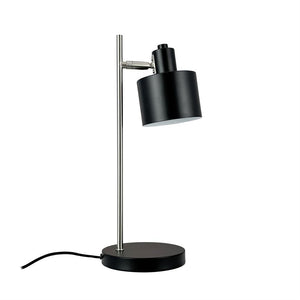 Skrivebordslampe med sort cylindrisk skærm, vipbar hoved, monteret på en slank sølvstang og rundt sort fod; sort ledning ligger ud til siden på hvid baggrund.