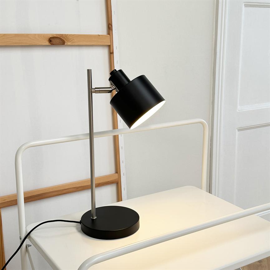 Sort skrivebordslampe lyser nedad, placeret på et hvidt bord foran en lys væg med en synlig træramme og en hvid dør i baggrunden.