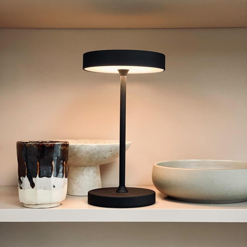 Bordlampe med slankt sort stativ lyser blødt varmt lys nedad; står midt på en lys hylde mellem en glasurbelagt keramikkop og to afdæmpede stenfarvede skåle.