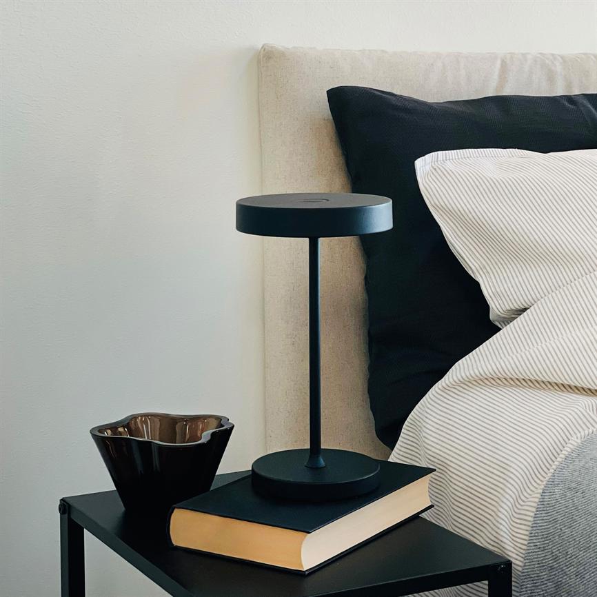 Sort moderne bordlampe står på en bog på et sort natbord; ved siden af en brun skål, bagved en seng med beige gavl, sort pude og stribet sengetøj.