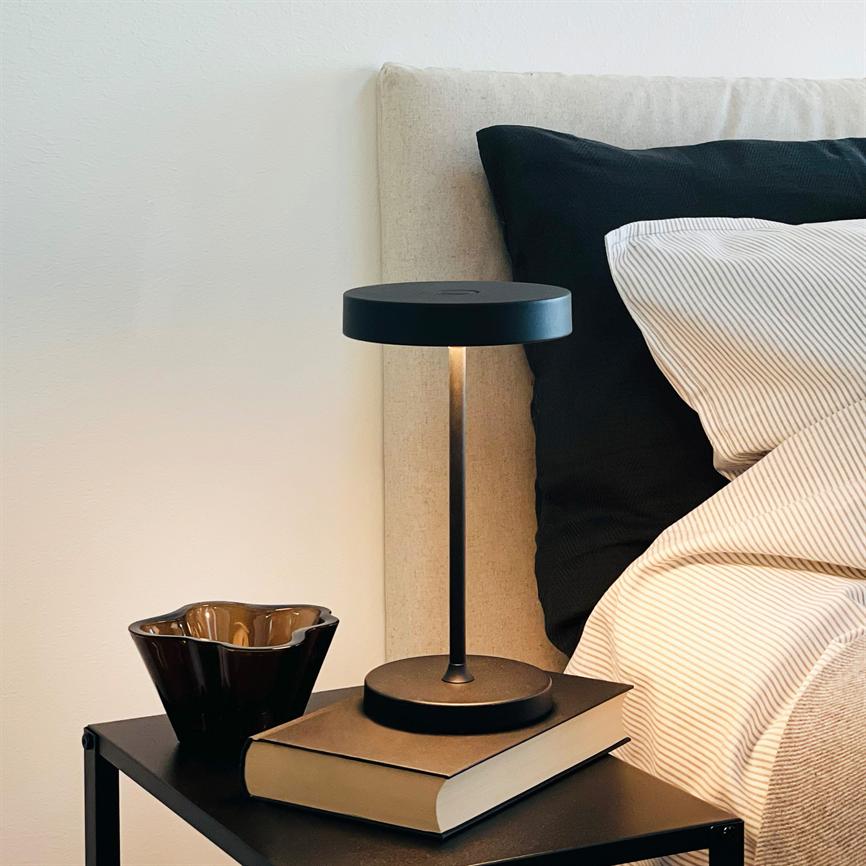 Bordlampe står tændt på en bog på et sort natbord; den oplyser et soveværelseshjørne ved en beige gavl og stribet sengetøj med sort pude, en brun skål ved siden.