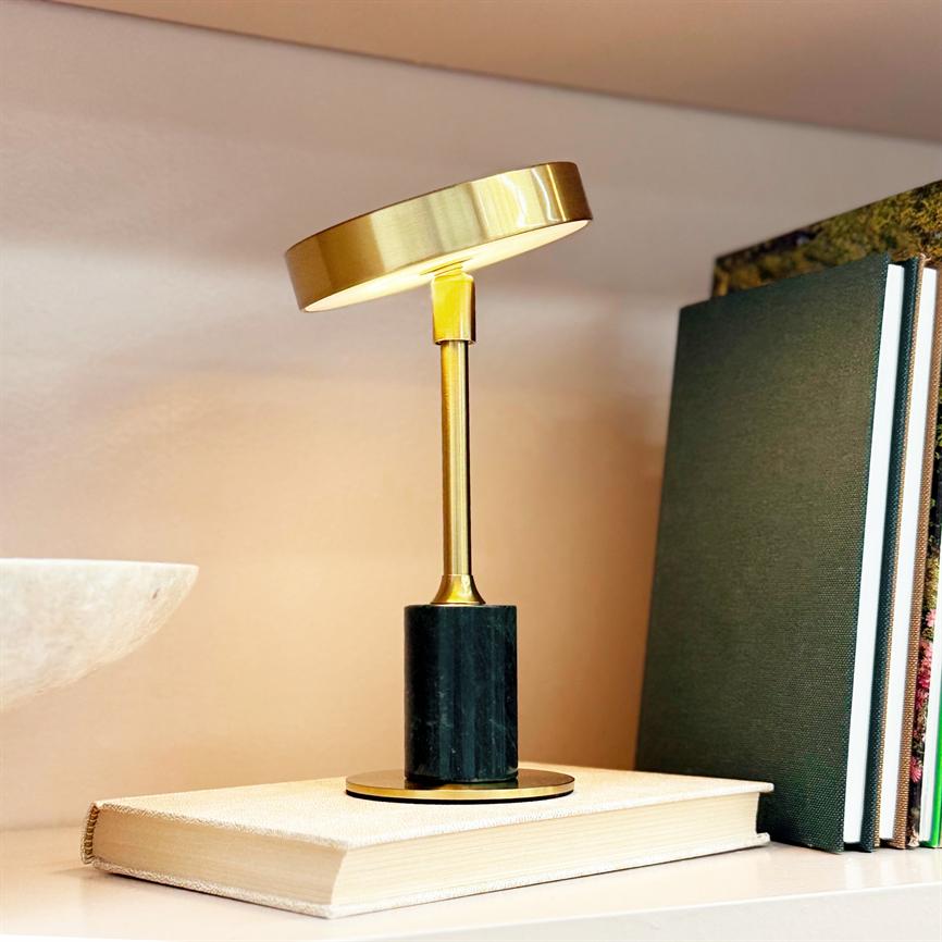 En lille guldfarvet bordlampe står på en lukket bog, kaster varmt lys; med sort marmorbase og stang, placeret på en hylde ved siden af mørke bøger og en hvid skål.