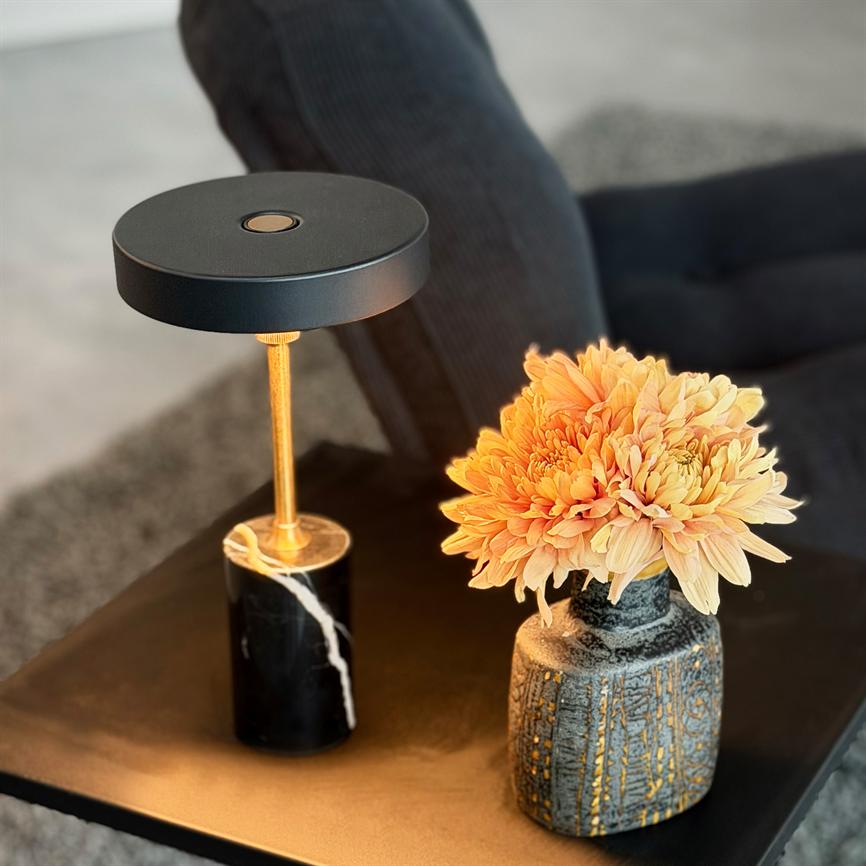 Bordlampen kaster varmt lys over en buket orange krysantemum i en keramisk vase; begge står på et sort sidebord foran en mørk sofa og et gråt tæppe.