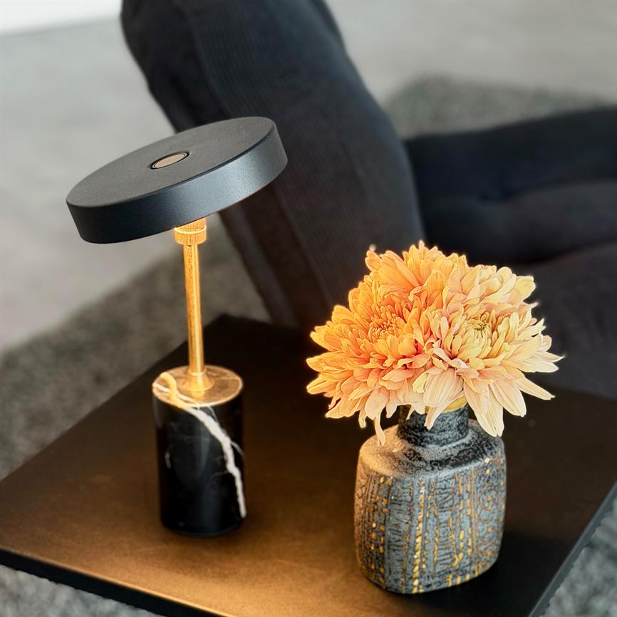 En buket lyse orange krysantemummer står i en keramisk vase; blomsterne oplyses blødt af en sort og guld bordlampe på et mørkt sofabord foran en lænestol.