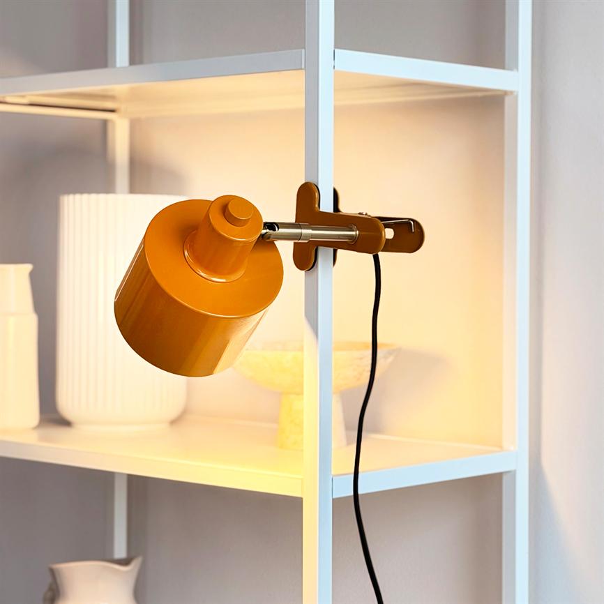 En orange klemme-lampe er fastgjort til en hvid metalreol og kaster varmt lys over hyldens hvide vaser og skåle; en sort ledning hænger ned.