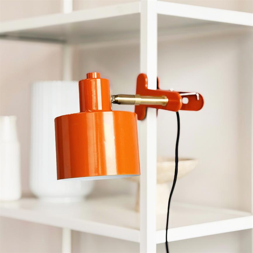 En orange metalvægslampe er fastklemmet til en hvid reol og peger nedad; en sort ledning hænger frit, omgivet af hvide vaser og neutrale dekorationer.