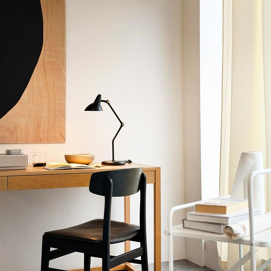 Sort vinklet skrivebordslampe lyser over en åben bog og en skål på et lyst træskrivebord; en sort træstol foran, i et solbeskinnet, minimalistisk hjørne ved vindue med beige gardin.
