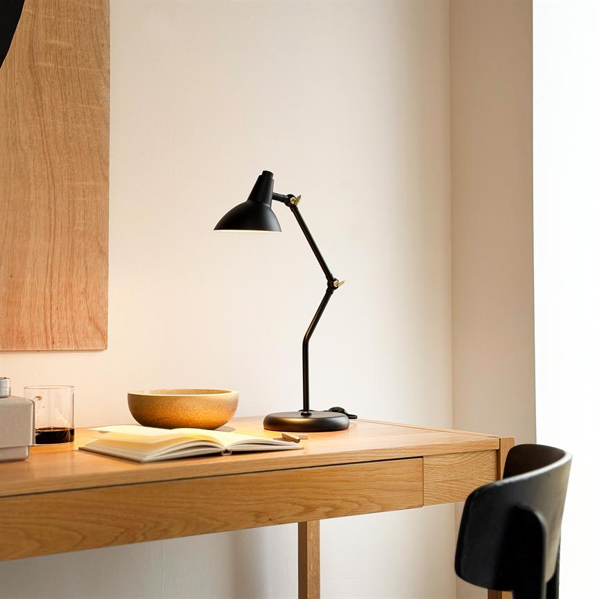 Sort, justerbar skrivebordslampe kaster varmt lys over en åben bog og en keramisk skål på et lyst træskrivebord i et enkelt, neutralt hjørne med en sort stol ved siden.