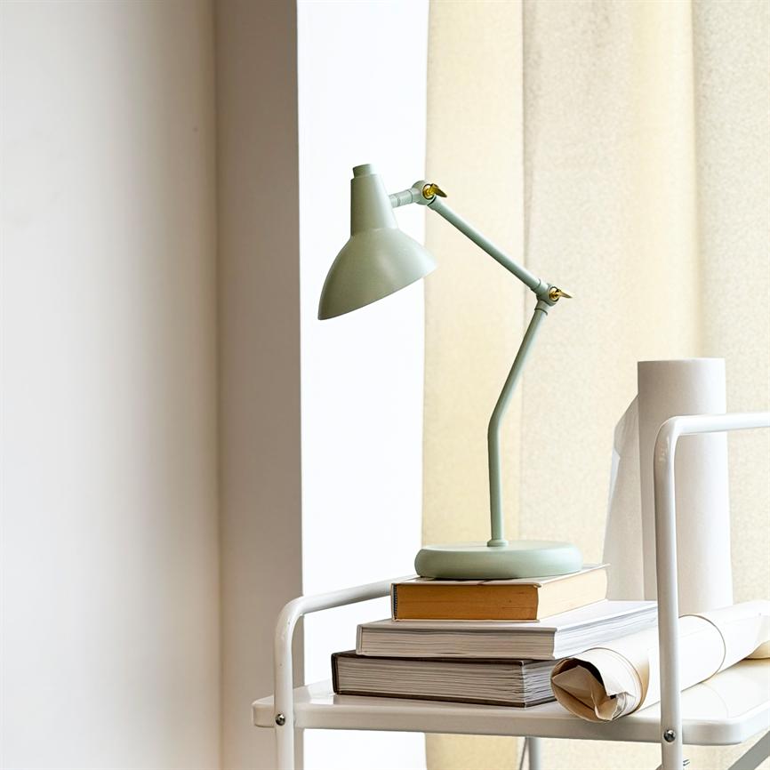 Grøn vinkelbar skrivebordslampe står på en stak bøger på en hvid rullevogn ved et vindue med lyse rullegardiner.