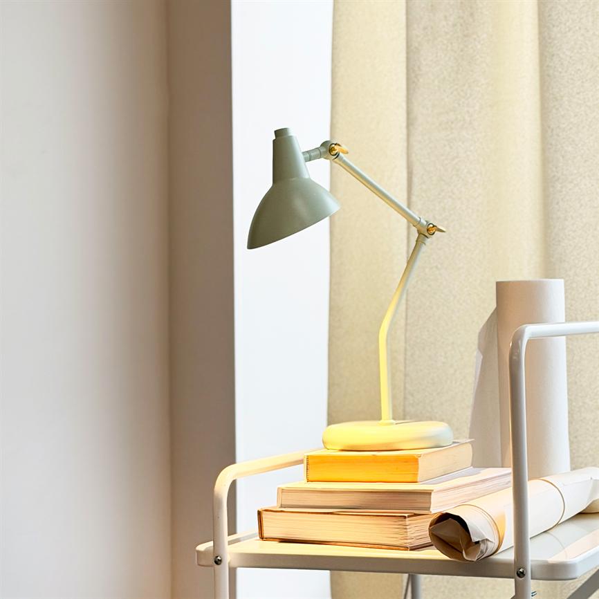 En grøn skrivebordslampe kaster varmt lys over en stak bøger på en hvid rullevogn ved et vindue med beige gardin og neutral væg bagved.