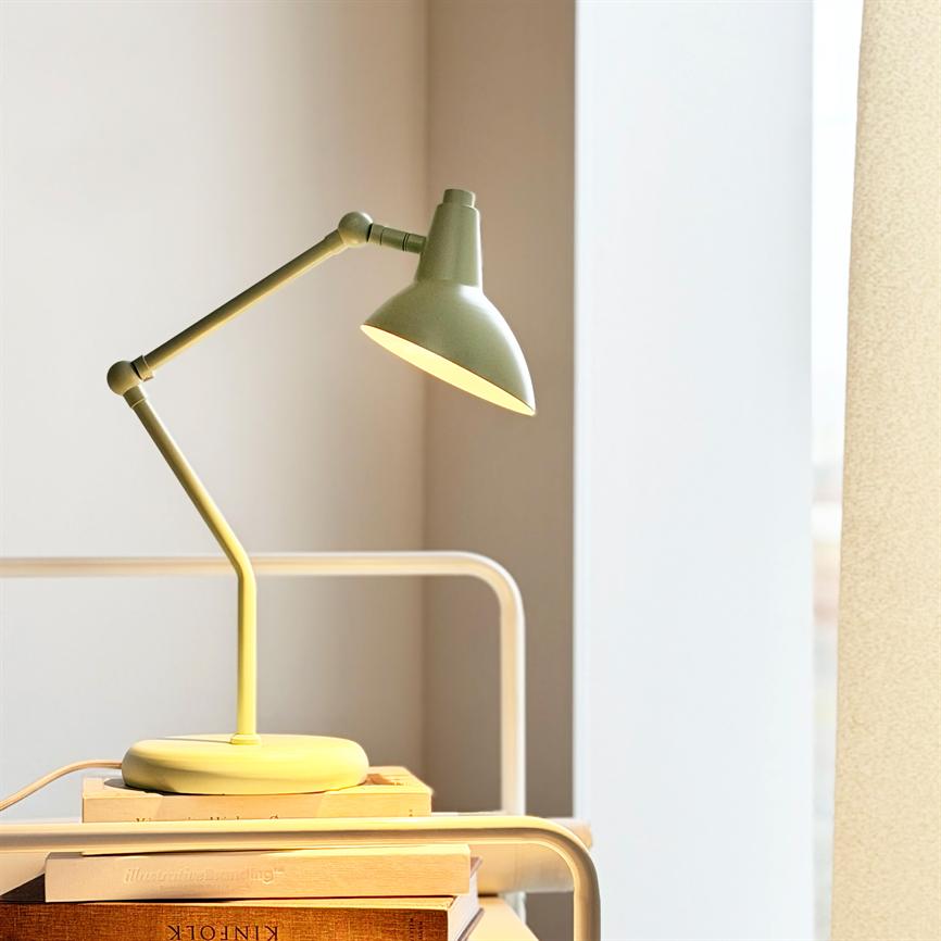 En olivengrøn skrivebordslampe lyser ned på en stak bøger; den står på et natbord ved en lys vindueskarm med et lyst gardin. Tekst: "KINFOLK".