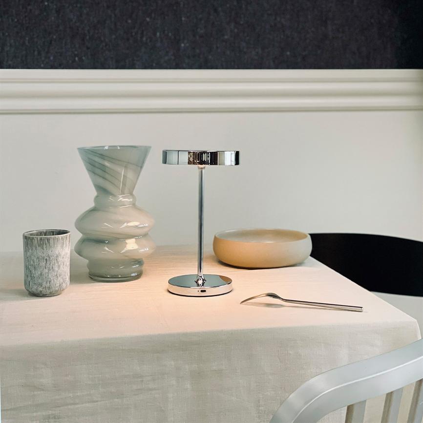 Krom bordlampe står tændt og kaster blødt lys over et hvidt, dugbetrukket bord; ved siden ligger en bølget glasvase, en lille kop, en rund skål og en ske.