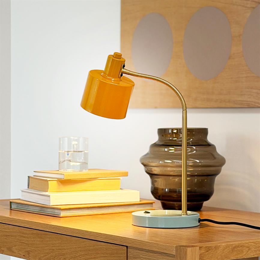 Gul skrivebordslampe med messingskaft er vinklet nedad og står på et træbord ved siden af en stak bøger, et glas vand og en mørk bølget glasvase i et lyst rum.