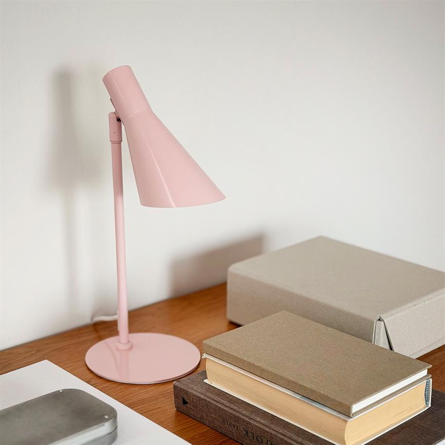 En lyserød skrivebordslampe står slukket og kaster en tydelig skygge; den står på et træbord ved siden af en stak bøger og en grå papæske mod en hvid væg.