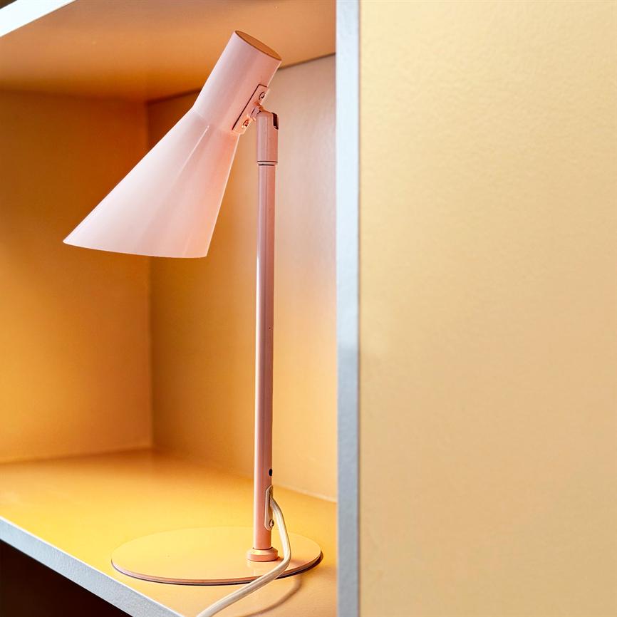 Bordlampe (lyserød) står tændt på en hylde i en indbygget niche, kaster varmt gult lys mod matte lysegule vægge, med hvid ledning snoet ved foden.
