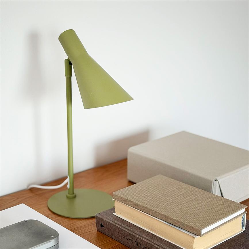 Grøn skrivebordslampe står ved siden af en stak bøger og en grå æske; lampen kaster en blød skygge mod en hvid væg på et træbord.