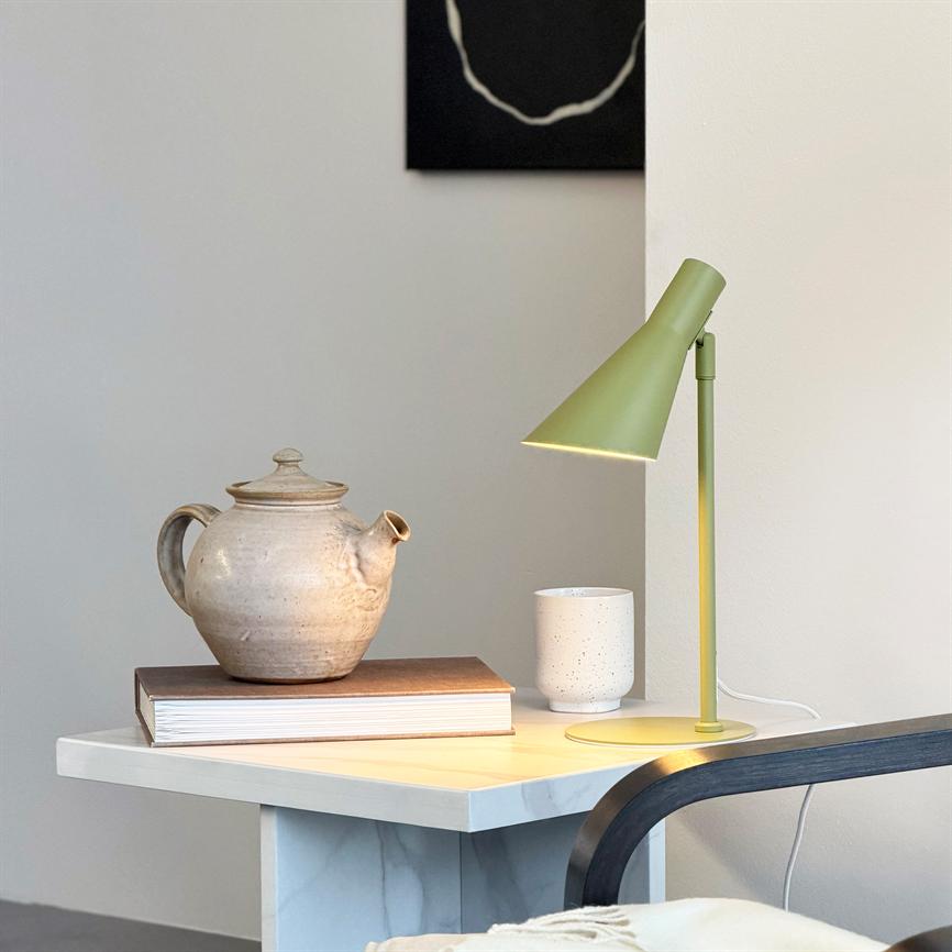 En keramisk tekande hviler på en bog, mens en grøn bordlampe kaster varmt lys over et marmor-natbord; en hvid krus og en stolarm er synlige mod en neutral væg.
