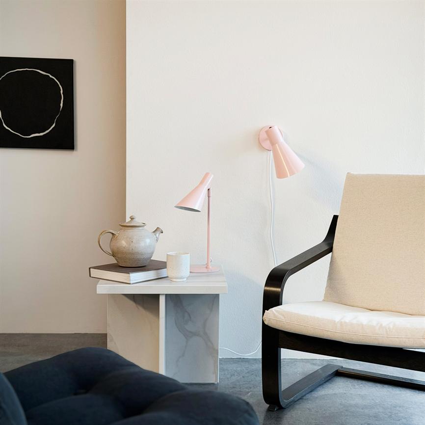 En beige lænestol står ved et lille marmorbord, hvor en tekande, en kop og en lyserød lampe er placeret; hvid væg og et sort abstrakt cirkelmaleri i et minimalistisk rum.