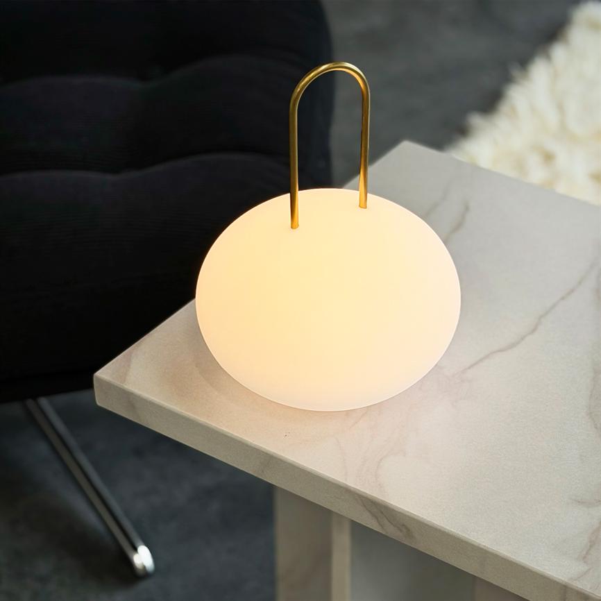 En rund, hvid bordlampe udsender varmt, diffust lys, hængt i et buet gyldent håndtag og placeret på hjørnet af et lyst, marmoreret sidebord ved en mørk lænestol.