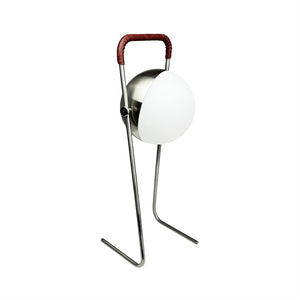 Bordlampe står på slanke metalben; rund hvid skærm fastholdt i børstet metal og buet håndtag med brunt læder, isoleret på hvid baggrund.