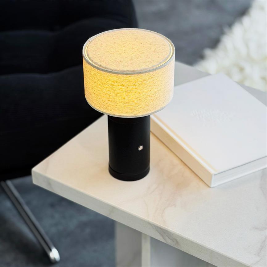 En bordlampe med sort cylindrisk fod og rundt lyst stofskærm udsender varm, diffus belysning; står på et lyst marmorbord ved en hvid bog og en mørk sofa i stuen.